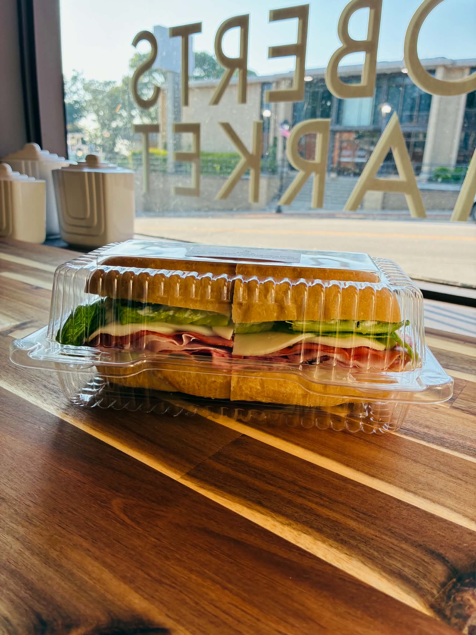 Grab-n-go sub with ham, salami, provolone, lettuce and tomato in a to-go container on a wood table by the window